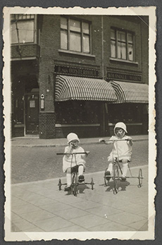 Thea en Leny op hun driewielers vóór de kruidenierswinkel van hun vader Jan van Oosterhout aan de Haagweg